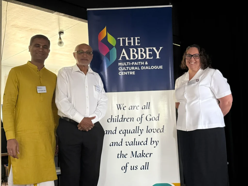 Three faith community representatives beside the Abbey multi faith centre's Statement of Belief banner, "We are all children of God and equally loved and valued by the Maker of us all"